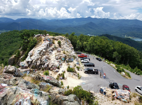 A scenic view from a rocky overlook, featuring a parking area with cars and mountains in the background under a cloudy sky.