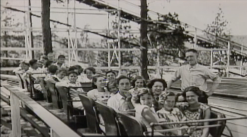 A vintage photo of a roller coaster with a group of smiling people seated in the cars, surrounded by trees.