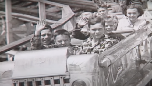 Black and white photo of children on a roller coaster, smiling and waving at the camera.