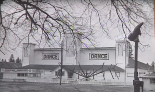 Black and white image of a building with "DANCE" signs, surrounded by trees and a streetlamp.