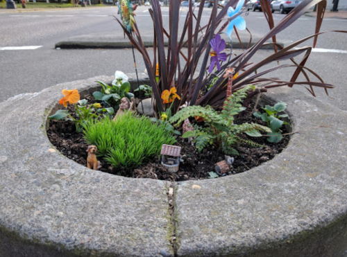 A small garden in a stone planter featuring flowers, ferns, and miniature decorations, including a dog and a house.
