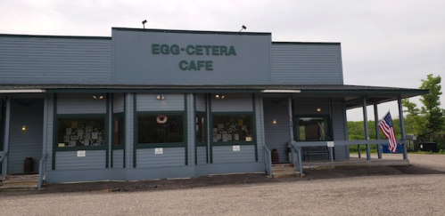Exterior of Egg-Cetera Cafe, a blue building with large windows and an American flag, set in a rural area.
