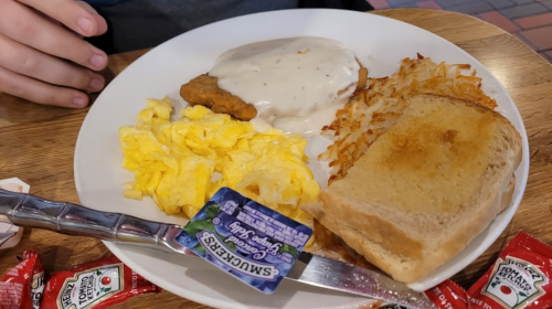 A plate with scrambled eggs, hash browns, chicken fried steak with gravy, toast, and a packet of jelly.