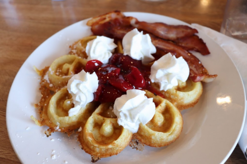 A plate of waffles topped with whipped cream and strawberry sauce, accompanied by crispy bacon strips.