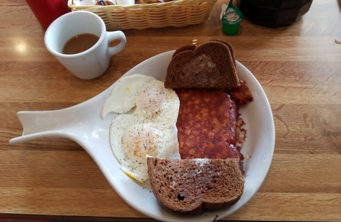 A unique plate shaped like a fish holds two fried eggs, a slice of cornbread, and two pieces of brown bread, with coffee.