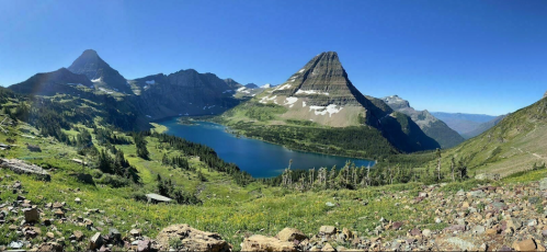 A panoramic view of a serene lake surrounded by mountains and lush greenery under a clear blue sky.