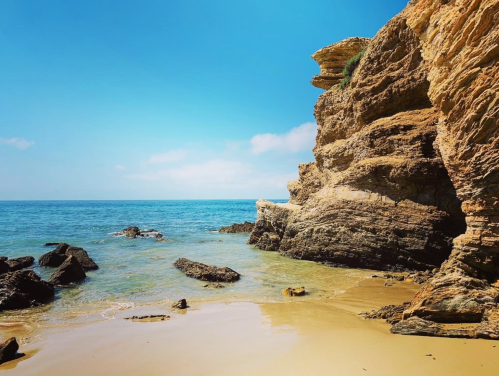 A serene beach scene with golden sand, rocky cliffs, and calm blue waters under a clear sky.