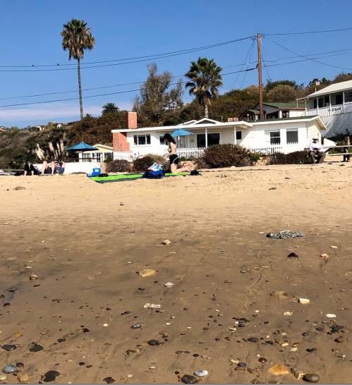 A sandy beach scene with people relaxing, palm trees, and beach houses in the background under a clear blue sky.