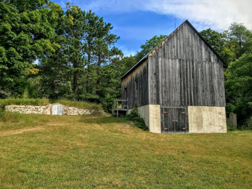 A rustic wooden barn beside a stone foundation, surrounded by green grass and trees under a blue sky.