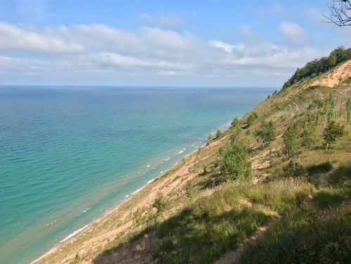 A scenic view of a coastline with a sandy slope, lush greenery, and calm blue waters under a partly cloudy sky.