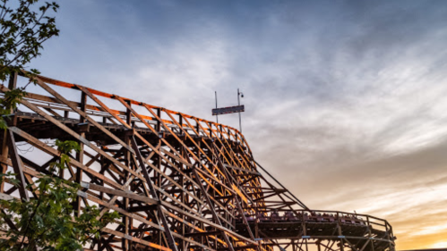 A wooden roller coaster structure against a sunset sky, with trees in the foreground.