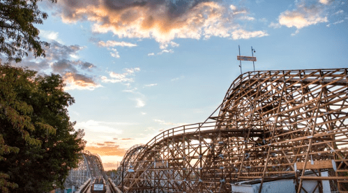A wooden roller coaster against a sunset sky with clouds, surrounded by trees.