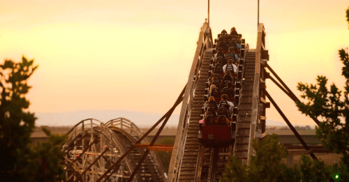 A roller coaster climbs a steep track at sunset, with riders visible in the cars against a colorful sky.