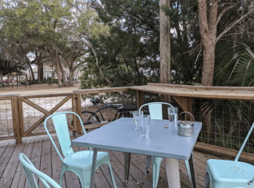 A wooden deck with a gray table and teal chairs, surrounded by trees and a glimpse of a bike in the background.
