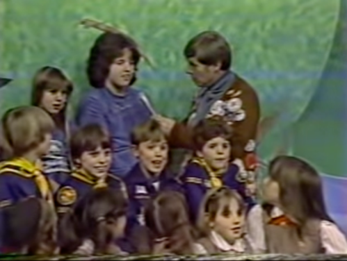 A group of children in uniforms sits with two adults on a colorful set, engaging in conversation and smiling.