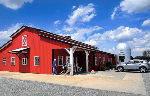 A red barn-style building with a white porch, people walking by, and a large silver dome structure nearby under a blue sky.