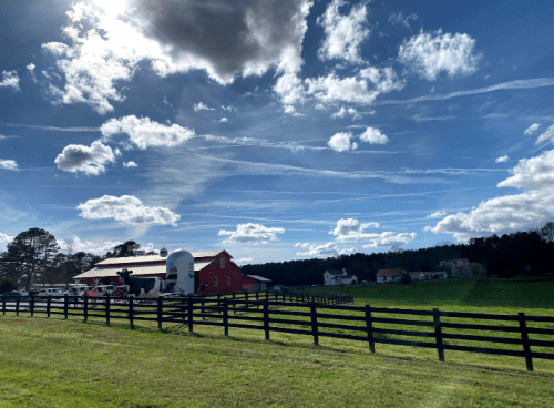 A red barn sits in a green field under a blue sky with fluffy clouds and wispy contrails. Fenced pasture in the foreground.