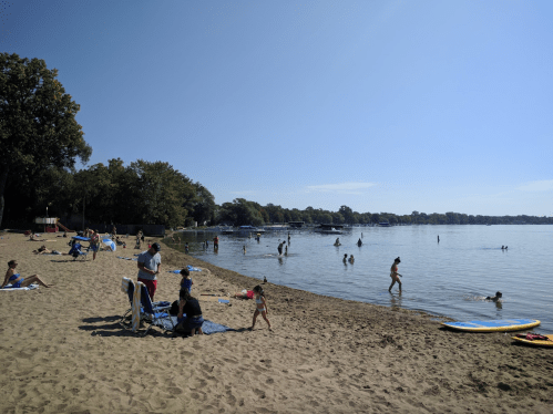 A sunny beach scene with people swimming, playing, and relaxing on the sand near the water's edge.