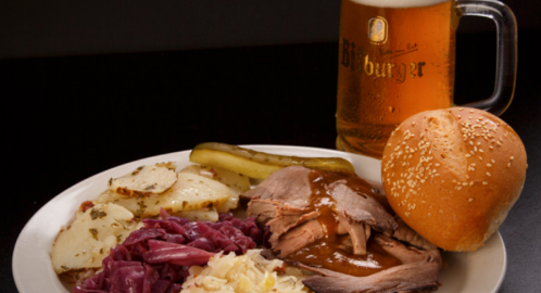 A plate of sliced roast beef with red cabbage, potatoes, pickles, and a sesame seed bun, alongside a glass of beer.