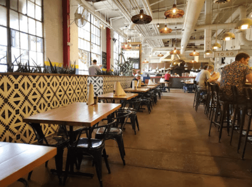A spacious restaurant interior with wooden tables, black chairs, and decorative walls, featuring diners and plants.