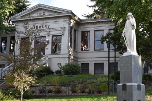 A historic library building from 1912, featuring a statue in front and surrounded by greenery.
