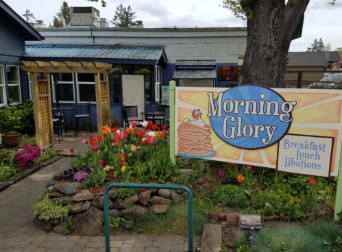 A colorful outdoor dining area with flowers and a sign for "Morning Glory" restaurant, offering breakfast and lunch.