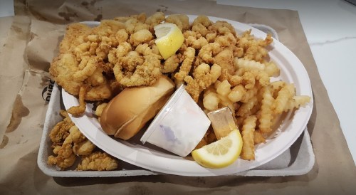 A large platter of fried seafood, including shrimp and fish, with crinkle-cut fries, a bun, and lemon wedges.