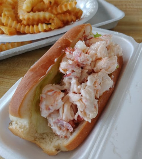 A lobster roll filled with lobster meat and lettuce, served in a bun, with crinkle-cut fries in the background.