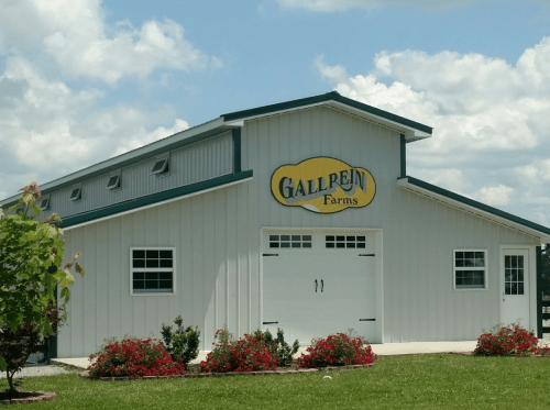 A white barn with a green roof and "Gallrein Farms" sign, surrounded by colorful flowers and a blue sky.