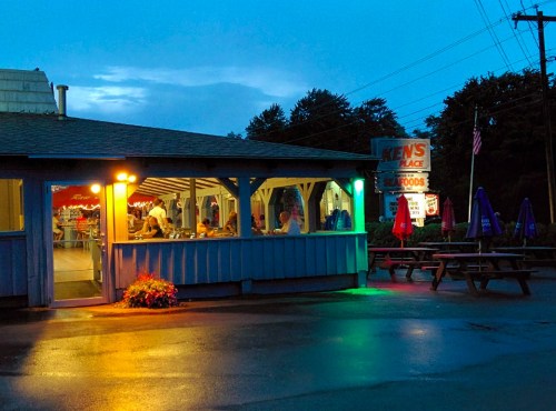 A cozy restaurant at dusk, with outdoor seating and colorful lights illuminating the entrance.