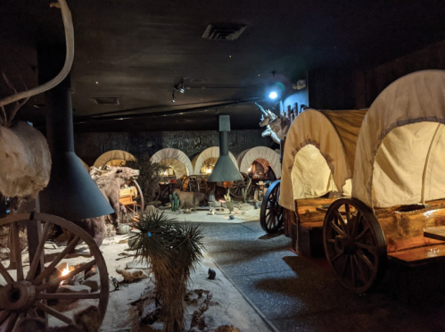 Interior of a dimly lit museum exhibit featuring covered wagons and various historical artifacts.