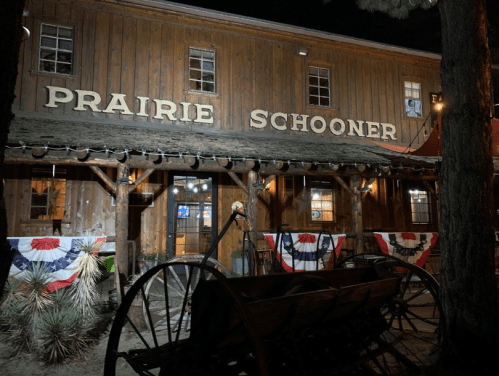 A rustic wooden building with "Prairie Schooner" sign, decorated with red, white, and blue bunting at night.