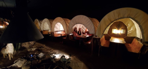 A cozy, dimly lit dining area with covered wagon-style booths and a campfire setup, creating a rustic atmosphere.