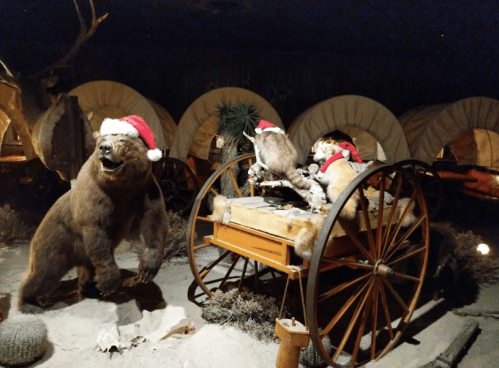A bear and two owls wearing Santa hats are near a wooden cart in a dimly lit, festive display.