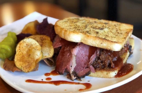 A hearty sandwich with layers of meat, served with potato chips and pickles on a white plate.