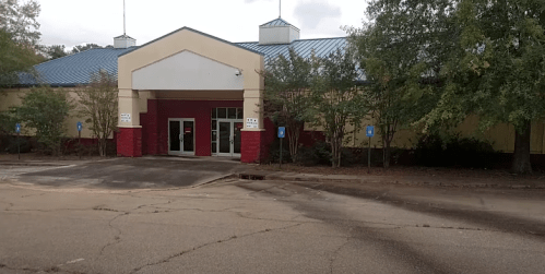 Exterior view of a building with a red and beige facade, a blue roof, and surrounding trees, set on a cracked parking lot.