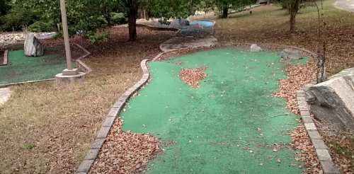 A mini-golf course with a green surface covered in fallen leaves, surrounded by trees and a winding path.