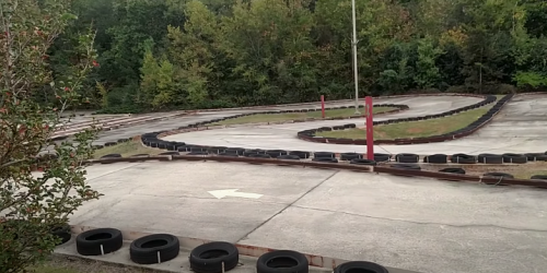 A go-kart track with a winding path, surrounded by trees and lined with tires, under a clear sky.