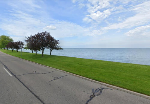 A serene lakeside view with a road, green grass, and trees under a partly cloudy sky.