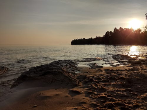 A serene beach scene at sunset, with calm water, rocky shore, and trees silhouetted against the glowing horizon.