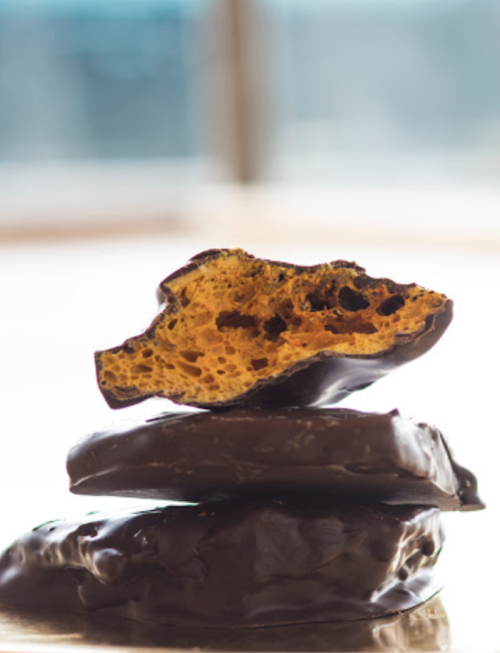 A close-up of a honeycomb candy piece partially covered in dark chocolate, stacked on two whole pieces.