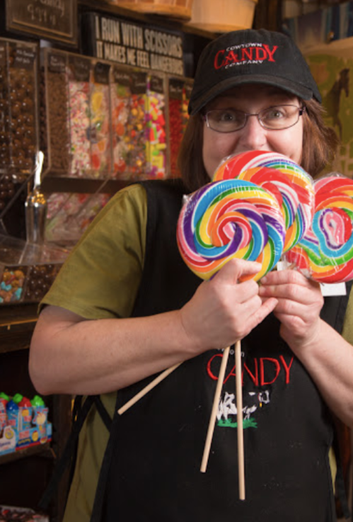 A smiling person in a candy shop holds colorful lollipops, surrounded by jars of sweets in the background.