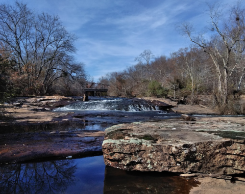 A serene landscape featuring a waterfall, rocky foreground, and bare trees under a blue sky.