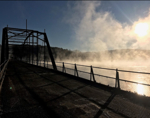 A misty river scene with a metal bridge silhouetted against the sun, creating a serene and atmospheric landscape.