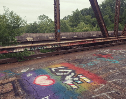 Colorful graffiti on a bridge, featuring a rainbow heart and the letters "SM," surrounded by greenery.