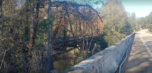 A rusted metal bridge surrounded by trees, with a road running alongside and a creek below.