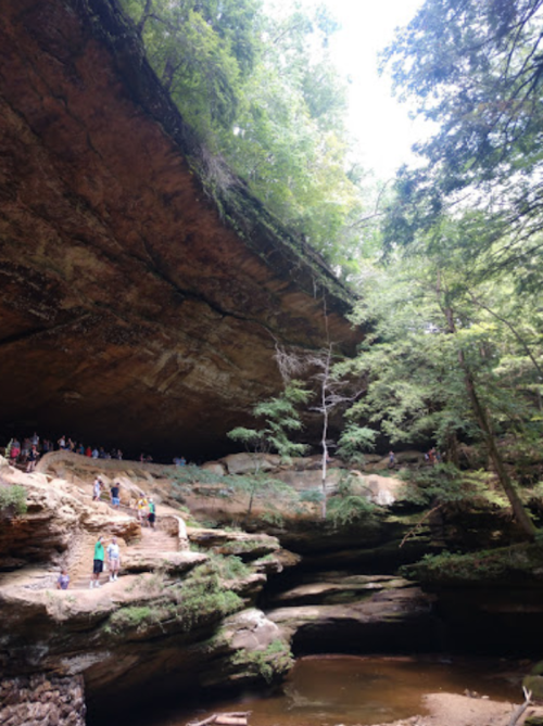 A scenic view of a rocky cave with lush greenery and people exploring the area near a calm water body.