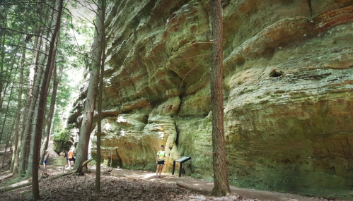 Hikers stand near a large, colorful rock formation in a forested area, with informational signs visible.