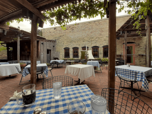 Outdoor dining area with checkered tablecloths, brick walls, and greenery overhead. Empty tables set for guests.