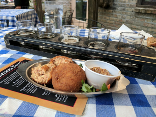 A metal plate with fried food and mustard, alongside a flight of beer glasses on a checkered tablecloth.